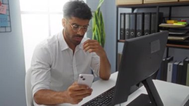 Akıllı telefon kullanan ve ofiste bilgisayarla uğraşırken stresli görünen genç bir adam.