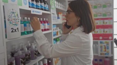 Eczanede beyaz laboratuvar önlüğü ve gözlük takan genç, çekici, İspanyol bir kadın. Dükkanın içindeki bir rafta ürünleri düzenlerken telefonla konuşuyor..