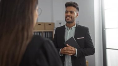 Ofiste kadınla konuşan erkek, elinde tablet tutarken arka planda raflar ve kutularla profesyonel giysiler içinde gülümsüyor..