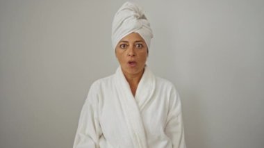 Bornoz ve havlu giymiş, orta yaşlı, şüpheli yüzlü, ağzı açık, alaycı ifadeli, beyaz arka planda soyutlanmış bir kadın.