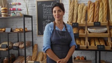 Bir fırında gülümseyen orta yaşlı bir kadın. Arka planda ekmek ve pastalar var. Kollarını kavuşturmuş, mavi gömlek ve önlük giyiyor..