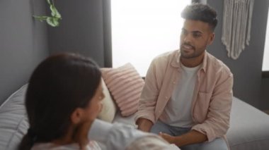 Kadın ve erkek, modern bir oturma odasında ciddi bir konuşma yapıyorlar. Pencereden içeri ışık giriyor.