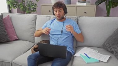 Evde çalışan rahat bir adam, yanında bir kediyle kanepede oturuyor, kulaklık takıyor, dizüstü bilgisayar kullanıyor ve rahat bir oturma odasında dinleniyor..