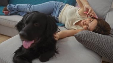 Genç bir kadın, rahat bir oturma odasında telefonla konuşurken siyah labrador köpeğiyle koltukta dinleniyor..