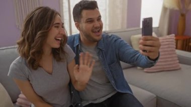 Gri koltukta oturan gülümseyen çift rahat bir oturma odasında, dekoratif yastıklar ve yumuşak ışıklandırmayla video görüşmesinin keyfini çıkarıyorlar.