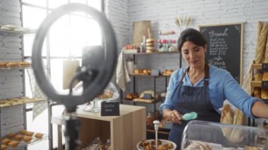 Kadın, sosyal medya için video çekerken pastanede hamur işleri hazırlıyor, çeşitli ekmek ve fırın ürünleriyle çevrili glütensiz bir tabela sergiliyor..