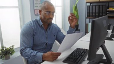 Ofiste çalışan, bilgisayarlı bir masada evrak okuyan, gözlüklü ve mavi gömlekli genç bir adam..