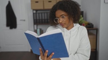 Kıvırcık saçlı ve gözlüklü Afro-Amerikalı genç bir kadın ofiste mavi bir kitap okuyor..