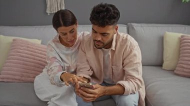 Oturma odasındaki kanepede oturan çift, birlikte akıllı telefona dikkatle bakıyor, ekranı işaret eden kadın, her ikisi de günlük kıyafetler giyiyor.