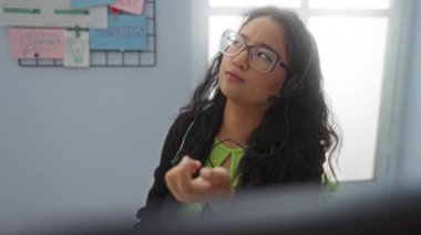 Ofiste kulaklıkla çalışan, gözlüklü ve yeşil gömlekli, arkasında strateji tahtası olan bir kadın..