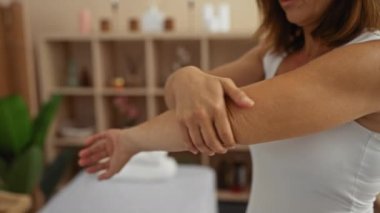 Orta yaşlı bir kadın kollarına losyon sürüyor. Kapalı bir kaplıca ortamında, kendine bakım ve cilt bakımını ön plana çıkarıyor.