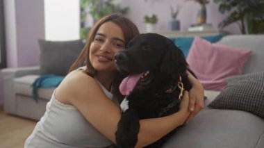Genç bir kadın, sıcak oturma odasında siyah köpeğe sarılıyor sıcak bir evde sevgi dolu bir bağ sergiliyor..