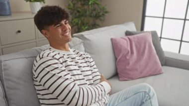 Modern dekorlu rahat bir oturma odasında rahat bir kanepede otururken gülümseyen İspanyol genç adam..