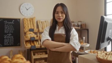 Fırında kendine güvenen bir kadın, kolları çapraz bir şekilde duruyor. Etrafı taze ekmek ve hamur işleriyle çevrili ve arkasında duvar saati olan bir yazar kasa kullanıyor.