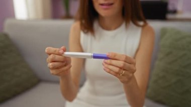 Elinde hamilelik testi olan genç bir kadın yeşil yastıklı ve bulanık arkaplanlı bir şekilde oturma odasında oturuyor.