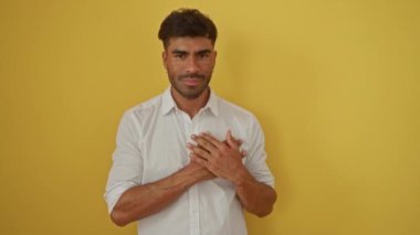 Elleri göğsünde çapraz duran genç adam, minnettarlığını gösteriyor, beyaz gömlekli, parlak sarı bir arka plana karşı duruyor..