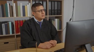 Yargıç cüppeli genç İspanyol adam arkasında kitaplıklarla bir ofiste çalışıyor.