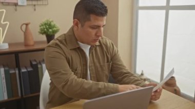 Evinde bir dizüstü bilgisayar üzerinde çalışan odaklanmış genç bir İspanyol. Evrakları ve modern dekorları var.