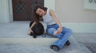 Şehir caddesinde oturan genç bir kadın dışarı çıkıyor, siyah labrador köpeğiyle selfie çekiyor, şehir yaşamını, bağlantısını ve arkadaşlığını vurguluyor..