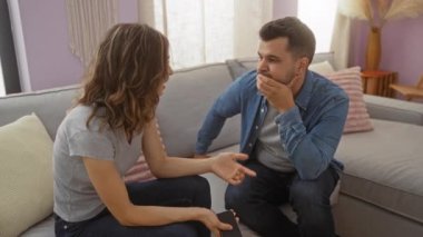 Modern bir oturma odasındaki koltukta konuşan bir çift, erkek dikkatle dinlerken telefonu tutan bir kadın, evde samimi bir konuşma hissi yaratıyor.