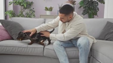 İspanyol adam evdeki modern oturma odasında kanepede dachshund okşuyor..
