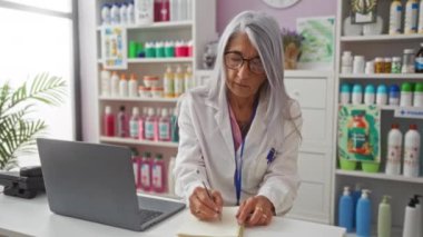 Gri saçlı yaşlı kadın eczacı not alıyor ve bir eczanenin içindeki dizüstü bilgisayarda çalışıyor. Etrafı çeşitli sağlık ürünleri ve ilaçlarla çevrili..