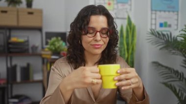Güzel, Latin kökenli, esmer bir kadın ofis ortamında, gözlük takıyor ve sarı bardaktan kahve yudumluyor..