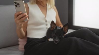 Oturma odasında kucağında chihuahua köpeğiyle oturan akıllı telefonu tutan bir kadın.