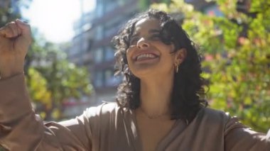 Neşeli, genç, esmer bir kadın güneşli bir günde yeşil ve binalarla çevrili şehir parkında manzaranın tadını çıkarıyor, mutluluk ve pozitifliği ifade ediyor..