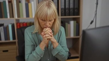 Ofisteki çalışma odasında kitap rafları ve bilgisayar monitörleriyle düşünen bir kadın, orta yaşlı, beyaz tenli bir kadını kapalı bir iş ortamında gösteriyor.