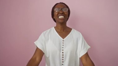 Gözlüklü bir kadın, çılgın bir ifadeyle duran genç bir Afrikalı Amerikalı kadın, izole edilmiş pembe arka plan yüzünden kızgın bir şekilde kollarını kaldırmış bağırıyor.