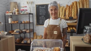 Bir pastanede çalışan yaşlı kadın neşeli tavırlı, kapalı bir ortamda taze ekmek ve hamur işleri sergiliyor.