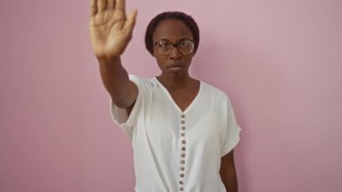 Gözlüklü genç Afro-Amerikalı bir kadın pembe arka planda duruyor. El hareketi ve yüzünde ciddi bir uyarı ifadesi var.