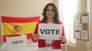 Genç bir İspanyol kadın, elinde bir İspanyol bayrağının önünde elinde bir oy levhası, seçim üniversitesi odasında gülümsüyor..