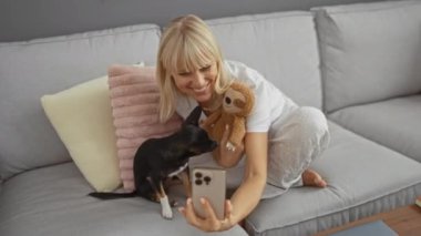 Orta yaşlı bir kadın Chihuahua köpeğiyle selfie çekiyor ve oturma odasındaki bir kanepede otururken oyuncak tutuyor.