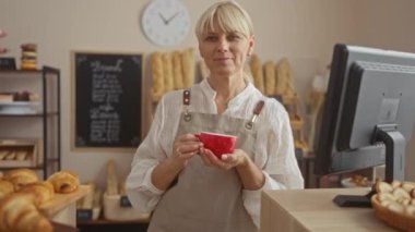 Fırında kırmızı kahve fincanı tutan olgun bir kadın fırıncı. İçi hamur işleri ve ekmek raflarıyla dolu.