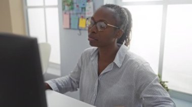 Ofiste çalışan Afrikalı Amerikalı bir kadın, bilgisayar ekranına odaklanıyor ve iyi aydınlatılmış bir iç mekan ortamında notlar alıyor..