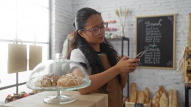 Fırında akıllı telefon kullanan bir kadın. Tezgahta karatahta menüsünün önünde hamur işleri sergileniyor.