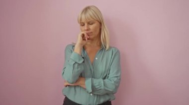 Kadın düşünen, orta yaşlı, sarışın, gömlek giyen, eli çenesinde, düşünceli ve ciddi bir yüz ifadesi olan, soyutlanmış pembe arka plan üzerinde düşünen ve soru soran bir kadın.