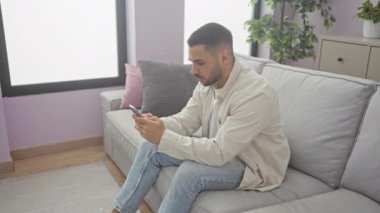 Yakışıklı İspanyol adam modern apartman dairesinde Dachshund 'la koltukta akıllı telefon kullanıyor.