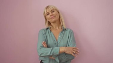 Mutlu orta yaşlı kadın, tişört giyen sarışın, çapraz kollu duran, gülümseyen ve pembe arka planda pozitif görünen.