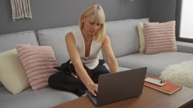 Oturma odasında laptopunu Chihuahua 'lı bir kadın kullanıyor. Ekrana bakıyor, eve odaklanmış.