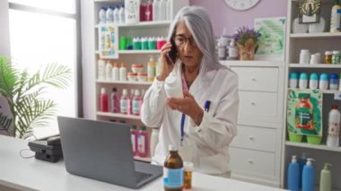 Laboratuvar önlüklü, gri saçlı yaşlı bir eczacı eczanede telefonla konuşuyor ve ilaç şişesini inceliyor.