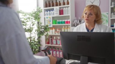 Yaşlı bir kadın eczacı, iyi organize edilmiş bir eczanenin içinde, raflardaki çeşitli tıbbi ve sağlık ürünleriyle çevrili bir müşteriye, tezgahtaki bilgisayarın arkasında yardımcı oluyor..