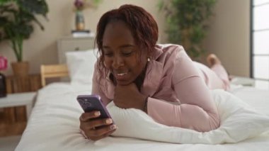 Pijamalı gülümseyen kadın, yatak odasında bitkilerle ve modern dekorlarla yatarken akıllı telefon kullanıyor.