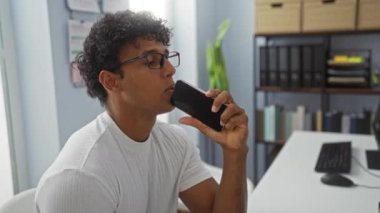 Modern ofisteki akıllı telefonu kullanan İspanyol genç adam, arka planda raflar ve bitkiler olan iyi ışıklandırılmış bir iş yerindeki cihaza yoğun bir şekilde odaklanıyor.