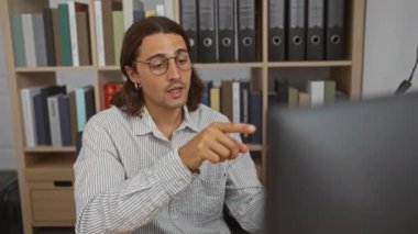 Ofiste çalışan uzun saçlı ve gözlüklü genç adam arkasında kitaplıklar olan bir bilgisayar ekranını işaret ediyor.