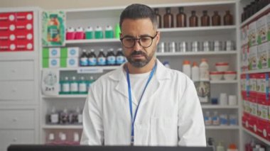 Eczacı üniformalı, ciddi, genç, İspanyol bir adam. Bir eczanenin içinde basit, doğal bir ifadeyle kameraya bakıyor.
