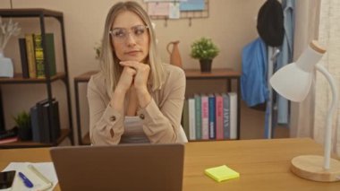 Sarışın, çekici, genç bir kadın rahat bir ofis odasında dizüstü bilgisayar üzerinde çalışıyor. Kitaplıklar ve arka planda saksı bitkisi var. Odaklanma ve tefekkür sergiliyor..