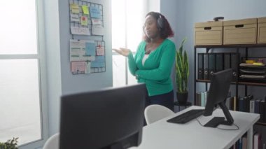 Genç Afro-Amerikalı kadın, modern bir ofiste, planlama tahtası ve bilgisayar ekranlarıyla el yapımı cihazla konuşuyor. Gündüz vakti içeride proje stratejisi üzerinde çalışıyor..